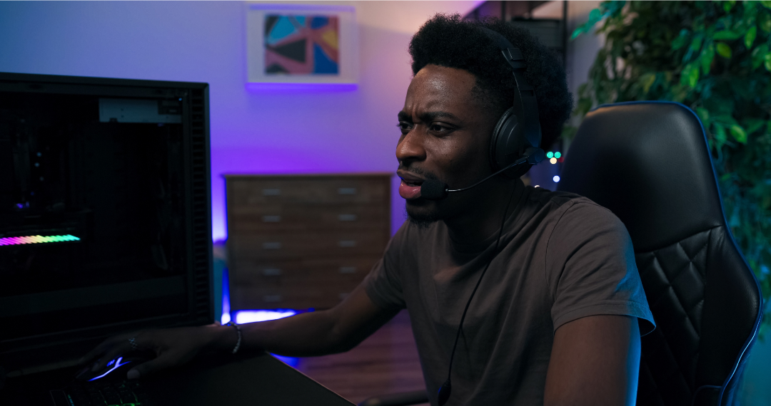 A man with a headset on is sitting at his gaming PC in a black office chair looking frustrated